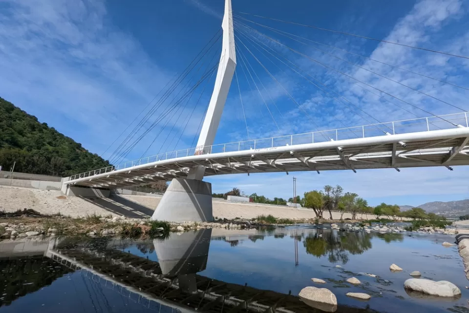 A cable-stayed bridge designed spans the water of the Los Angeles River, which is placid and dotted with rocks.